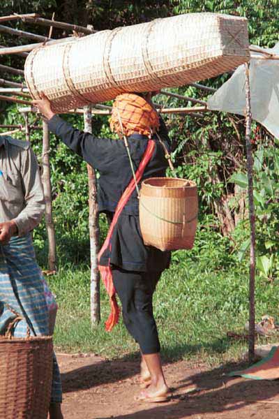 46K Jpeg 9809H17 Pa'O woman carrying her baskets for sale into Kalaw 5-day market, Shan State. Note her black serge three-layers of clothing - short jacket over aloose blouse (note the blue and red over-sewn embroidery on the side seam) with both worn over a calve length longyi. She has a finely woven basket on her back.