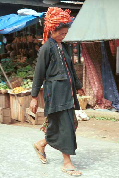 26K Jpeg 9809K01 Pa'O woman striding through the 5-day rotating market in Kalaw, Shan State. Her garments of black serge short, long-sleeved jacket with selvedge woven trim inset down the opening of the jacket, loose blouse with embroidered trim along the seams and calf length longyi are set off by the plaid scarf wound around her head and her striped bag