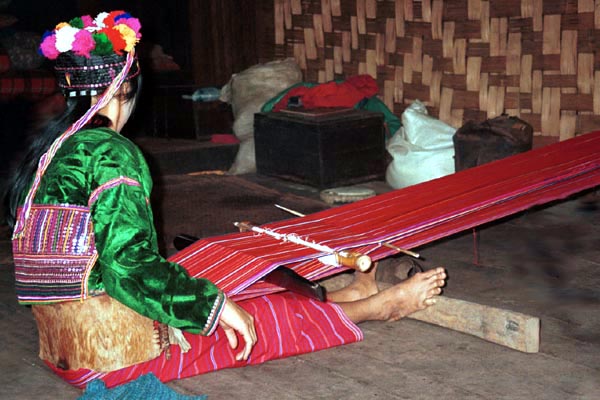 73K Jpeg Silver Palaung woman in Pein Ne Bin village, near Kalaw in southwestern Shan State, weaving a length of cotton fabric to make a tube skirt similar to the one she is wearing. Note the narrow stripes in warp threads. She is using a very simple backstrap loom with a wide leather back support. She is wearing a velvet decorated cap which is reputed to be worn by young girls and unmarried women although she appeared to be a married woman with a husband and baby. She has a very wide band of decoration using many rows of sequins on the back of her blouse.