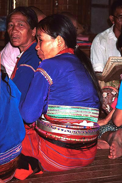 76K Jpeg Silver Palaung women in the Buddhist temple at Pein Ne Bin village near Kalaw, northwestern Shan State joining in the prayers on the day after the full moon 'Full Moon Day". They are wearing traditional hand woven tube skirts, embroidered blouses of a slightly shiny fabric and the Silver Palaung identifying bamboo or rattan hoops. The woman in the centre of the photo is also wearing a white metal belt. A white sash and black silky threads can also been seen amongst the hoops.