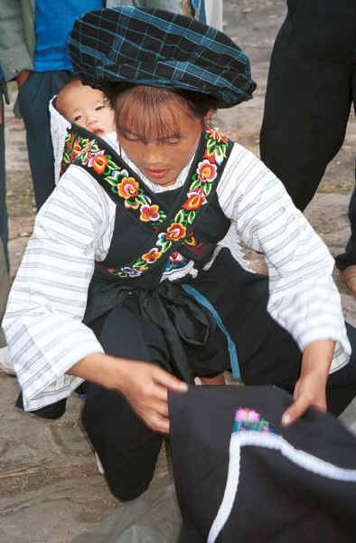 Bouyei mother with her baby on her back spreading out an apron for sale - Bi Ke village, Mi Gu township, Zhenfeng county, Guizhou province 0010s22.jpg