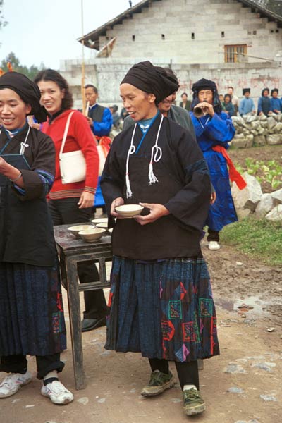 Jpeg 46K  0110D13 Women offering us alchohol as part of our welcome to Gan He village, Ya Rong township, Huishui county, Guizhou province, south-west China. The people living in this village are known as Qing Miao. There has been much intermarriage between Miao and Bouyei which is evident from the every-day costume of the women. The pieced skirts of applique and wax-resist were originally only worn at funerals but today they are worn for special festivals and dance competitions.