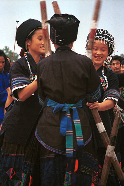 Jpeg 57K  0110D36 Girls beating sticky rice as part of a dance programme in Gan He village, Ya Rong township, Huishui county, Guizhou province, south-west China. The people living in this village are known as Qing Miao. There has been much intermarriage between Miao and Bouyei which is evident from the every-day costume of the women. The pieced skirts of applique and wax-resist being worn by the girls were originally only worn at funerals but today they are worn for special festivals and dance competitions. Note the bands of complex weaving on the sleeves of the women's jackets.