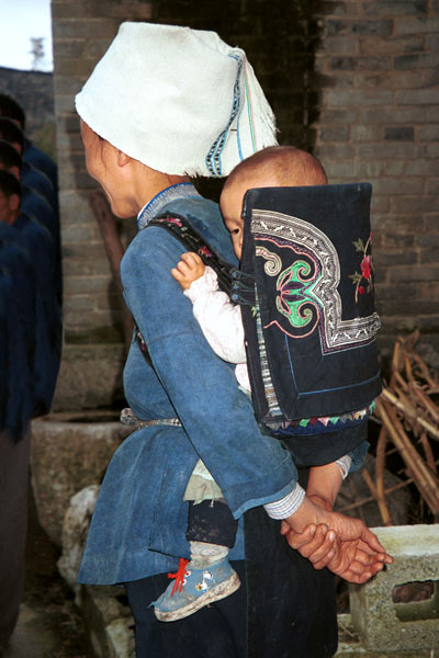 Jpeg 56K  0110E16 A baby held in an attractive baby carrier with embroidery and applied braid in Gan He village, Ya Rong township, Huishui county, Guizhou province. This is a Qing Miao (or Bouyei/Miao as there has been much intermarriage and the costume is very mixed). 