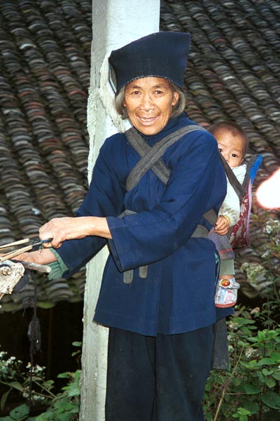 Jpeg 50K 0110E17  An old woman with a baby in a baby carrier on her back holding the ends of warp threads which are being untangled and straightened before warping up a loom in Gan He village, Ya Rong township, Huishui county, Guizhou province. This is a Qing Miao (or Bouyei/Miao as there has been much intermarriage and the costume is very mixed).