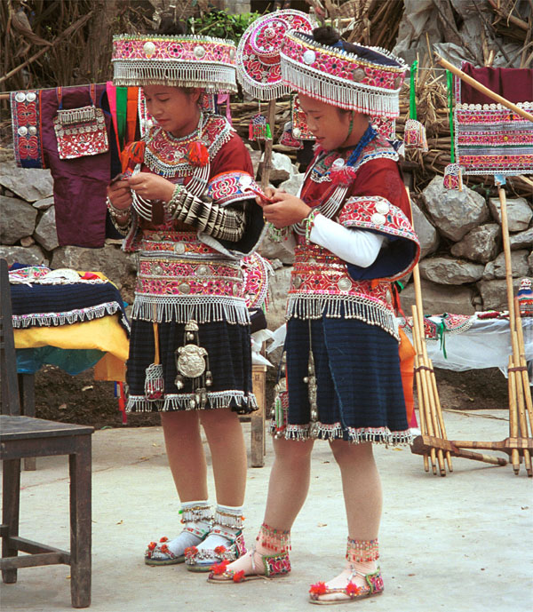 Jpeg 176 Two Iron beating Miao women in Gao Zhai village, Bai Jin township, Huishui county, Guizhou province working on some embroidery and showing off their festival costume against a back drop of an even brighter hat and two of their jackets 0110C09