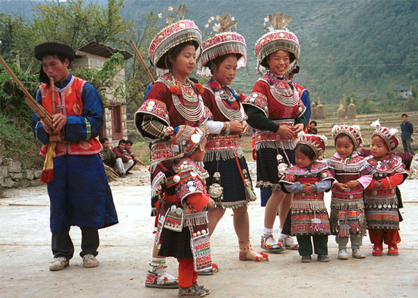 Jpeg 111K The welcoming dance troup of little Iron beating Miao girls, women and men with lusheng pipes against a beautiful backdrop of the rice harvest and tall haystacks agains the mountain background. These costumes will no doubt be used in dance competions as well as for festival occasions. Gao Zhai village, Bai Jin township, Huishui county, Guizhou province 0110C18A