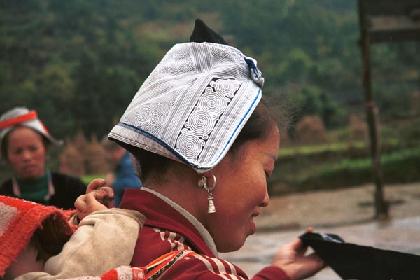 Jpeg 38K 0111F31 Gejia woman carrying her baby on her back and showing her headdress. The fabric is a machine printed reproduction of a traditional Gejia wax resist design. All the women in this village seemed to be wearing the commercially printed fabric and we did not see any original wax resist being worn on headdresses. Ma Tang village, Kaili City, Guizhou province. 