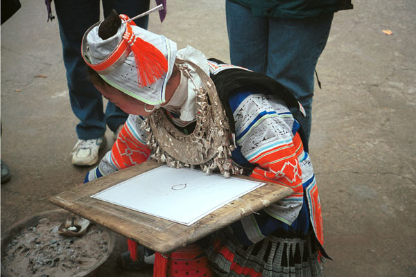 Jpeg 58K 0100F34 Gejia woman dressed in full festival finery demonstrating wax resist. The design on her own costume - her headdress and jacket at least (the black on white) - is probably all made from a commercially printed fabric and is not made from traditional wax resist. She has heated the wax in a metal dish on hot ashes. Ma Tang village, Kaili City, Guizhou province.