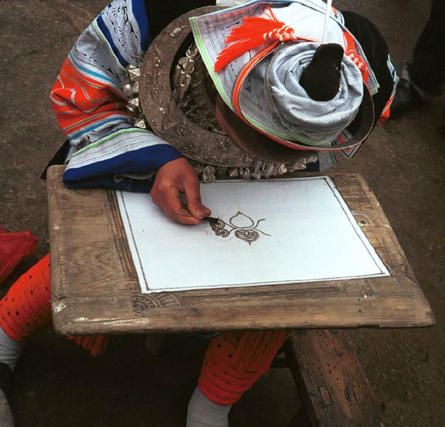 Jpeg 60K 0111G02 Gejia woman in full festival finery demonstating wax resist. Ironically all the apparently waxed material on her costum machine-made commercial fabric based on traditional Gejia wax resist designs. Ma Tang village, Kaili City, Guizhou province.