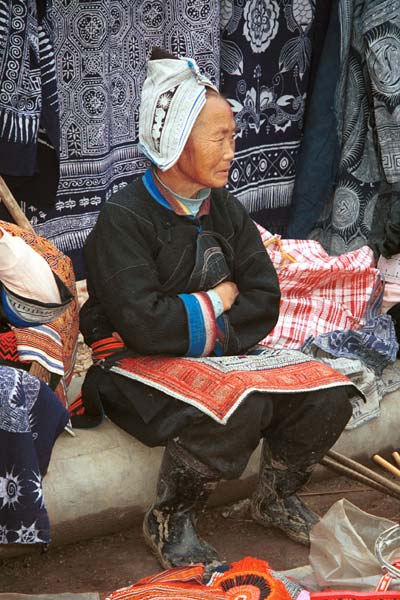 Jpeg 57K 0111G30 Gejia woman sitting amongs her modern wax resist work made to sell to the tourists. Note her embroidered apron which is edged with some original wax resist. Her headdress is, however, made from a commercially manufactred fabric using traditional Gejia designs. Ma Tang village, Kaili City, Guizhou