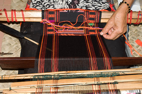 Weaver Huang Ji Xiang, who is a member of the Qi subgroup of the Li people, making the cloth that is called "Li brocade" locally. Weaving proceeds via two separate sheds. A lower shed is opened with the large, dark colored weaving sword, which is twisted to open the shed wider. Black weft is passed through on a spindle and then beaten down with the sword.