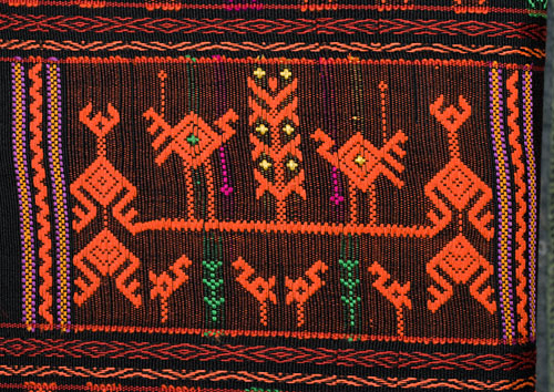 A design on a cloth is for the tourist market, rather gaudy and with large, widely spaced designs (for ease and speed of production I expect), but Chris thinks that the basic technique is the same used to make most Li minority supplementary weft weavings. Note in areas which are left without design the weft underneath tends to show through (see green threads in this design), which is somewhat inelegant, and may explain why good, old Li weavings tend to have very dense designs without much "ground" visible.