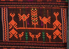 Jpg 161K - A design on a cloth is for the tourist market, rather gaudy and with large, widely spaced designs (for ease and speed of production I expect), but Chris thinks that the basic technique is the same used to make most Li minority supplementary weft weavings. Note in areas which are left without design the weft underneath tends to show through (see green threads in this design), which is somewhat inelegant, and may explain why good, old Li weavings tend to have very dense designs without much "ground" visible.