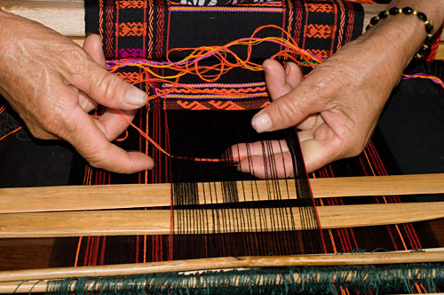 Weaver Huang Ji Xiang, who is a member of the Qi subgroup of the Li people, making the cloth that is called "Li brocade" locally. Weaving proceeds via two separate sheds. After adding a row of ground weft the weaver switches attention to the upper shed where the pattern is formed. She first selects the region of interest with the light colored wooden pick at the bottom of the photo, then uses a second pick to select the warps for the supplementary weft, which is then added by passing the thread from hand to hand because the distance is too short to need a spindle or shuttle. She then beats this down using the pick/sword and switches back to the lower shed ... and so the weaving goes.