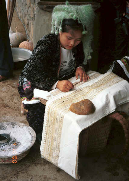 Young Side comb Miao woman sitting waxing the batik which will be indigo dyed to go at the top of a skirt.  She is copying the batik from another skirt and has a lump of bees wax in front of her on which she wipes excess wax off her waxing instrument and a pot of melted wax sitting in a bowl of hot embers beside her.  Long Dong village, De Wo township, Longlin country, Guangxi province 0010e21.jpg