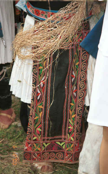 Front view of a White Miao woman showing her machine embroidered apron over a pleated hemp or ramie woven skirt over which is draped hemp or ramie fibre stripped from the bush which she is splicing as she stands and waits - Ma Wo village, Zhe Lang township, Longlin county, Guangxi province 0010j37.jpg