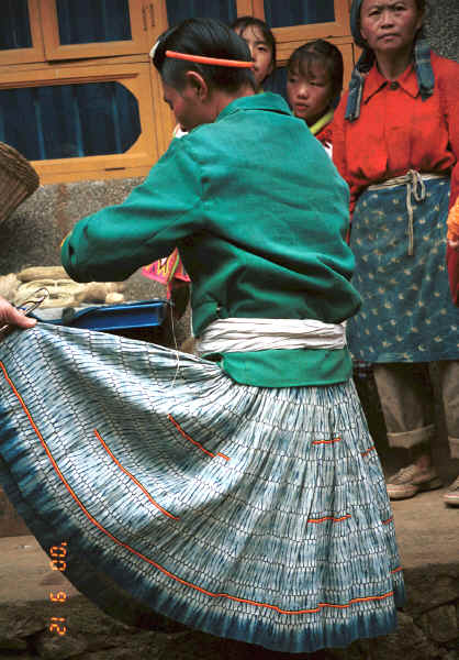 Big Flower Miao woman showing her skirt where the pattern has been created by indigo dye and stitch resist rather than batik as has the apron on the woman in the background.  This stitch resist is preferred over batik for work clothes as the women said that it was more hard wearing - it probably takes longer to do the batik wax resist which would be another reason for keeping it for more special occasions.   Xian Ma village, Hou Chang township, Puding county, Guizhou province 0010y05.jpg