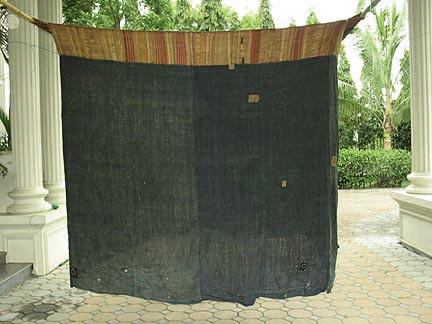 Jpeg 50K Tai Daeng Mosquito Net (pha mung) from Xam Tai in northeast Laos. Approximately: 61" x 65" on the sides and 60" high; hanging pockets extends 4" from main 'box'. Four panels on each side of handspun, handwoven indigo cotton, with silk and cotton heading border in good condition; top of four panels (one is pieced making three parts) gold-colored handspun, handwoven cotton. One side (side 'd') has a panel replaced, or of darker color indigo. Several patches and some holes, although none are larger than 1". A few patches of supplementary warp scraps. All handstitched. Early 20th century.