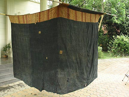 Jpeg 57K Tai Daeng Mosquito Net (pha mung) from Xam Tai in northeast Laos. Approximately: 61" x 65" on the sides and 60" high; hanging pockets extends 4" from main 'box'. Four panels on each side of handspun, handwoven indigo cotton, with silk and cotton heading border in good condition; top of four panels (one is pieced making three parts) gold-colored handspun, handwoven cotton. One side (side 'd') has a panel replaced, or of darker color indigo. Several patches and some holes, although none are larger than 1". A few patches of supplementary warp scraps. All handstitched. Early 20th century.