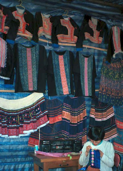 Blue Hmong woman sewing applique in front of an array of clothing 8812l16A