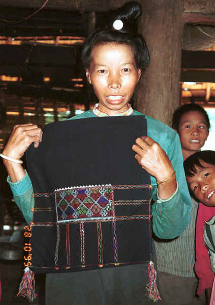 Jpeg 29K Black Thai woman showing a head cloth which she embroidered several years before - the style has less ornate edging than ones being made and worn today.  Note that, inside the house, she is not wearing a head cloth but showing her hair knot secured by a silver coloured coin fastening.  This is the lady who was demonstrating weaving in the loom above for which we went to a neighbour's house as this Black Thai lady did not have a loom currently set up in her house.  The lady of the house - shown in the photo to the right of this one with a more modern head cloth - was out collecting wood for her cooking fire when we first arrived at her home and watched the weaving.  Both head cloths are now in my collection purchased by way of thanks and in memory of these ladies' kind and generous hospitality.  Dien Bien Phu, Lai Chau Province. 9510D27.JPG