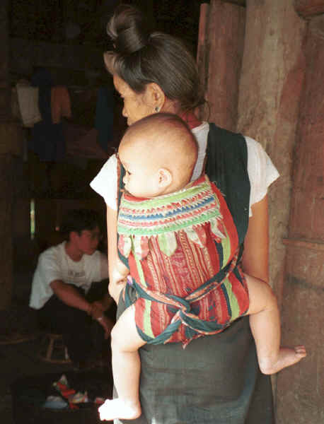 Jpeg 26K Black Thai baby being carried on it's grandmother's back in a baby carrier made with an insertion of supplementary woven cloth and layers of edging made from individually folded 'points'.  A village near Dien Bien Phu, Lai Chau Province. 9510E21.JPG