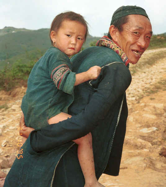 Jpeg 30K Black Hmong man with a young girl - probably his granddaughter - on his back, in the hills around Sa Pa, Lao Cai Province.  Note the beautifully embroidered collar back on his sleeveless indigo dyed jacket. 9510K13.JPG