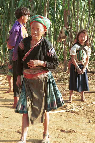 Jpeg 44 K  9510G2Older Flowery Hmong woman with children outside a house in small road-side village near Phong Tho (Phong Thanh) in Lai Chau Province. Note her short pleated skirt with batik and apliqued fabric squares and also her long, indigo apron.  Her head cloth will have been bought in the market - machine woven and imported from China.