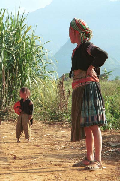 Jpeg 49K  9510G24 Older Flowery Hmong woman and child outside a house in small road-side village near Phong Tho (Phong Thanh) in Lai Chau Province. Note her short pleated skirt with batik and apliqued fabric squares and also her long, indigo apron.  Her head cloth will have been bought in the market - machine woven and imported from China