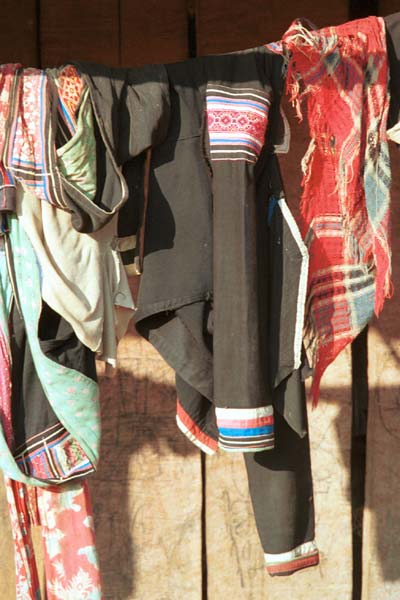 Jpeg 42K  9510G25 Flowery Hmong clothing hanging out to dry outside a house in small road-side village near Phong Tho (Phong Thanh) in Lai Chau Province. A woman's jacket with applied strips over the shoulders and at the end of the sleeves and with some embroidery.  To the left is probably the long waist tie and apron or possibly a baby carrier.  The head cloth will have been bought in the market - machine woven and imported from China.