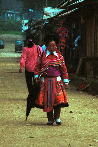 Jpeg 48K 9510K26  Brightly dressed Flowery Hmong woman walking down the street in Sa Pa, Lao Cai Province.  She is a visitor to Sa Pa, in the town on a training programme, and lives near the Chinese border. The Flowery Hmong near to the border tend to wear more bought trimmings on their clothing which they obtain from China.  Note her embroidered and applique leg wrappings and her long apron.  She only very narrow bands of wax resist on her skirt which features applied strips of fabric and embroidery. 