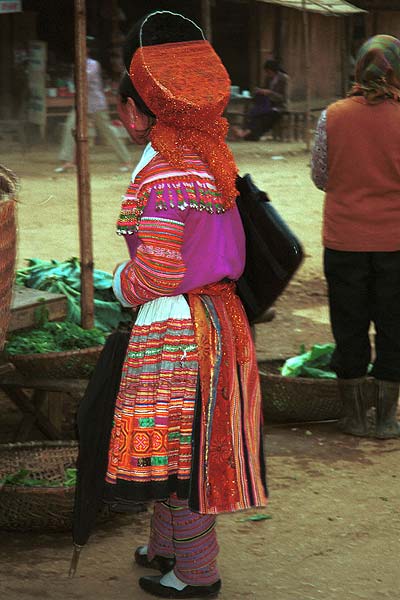 Jpeg 45K 9510K27  Brightly dressed Flowery Hmong woman at a shop stall in Sa Pa, Lao Cai Province.  She is a visitor to Sa Pa, in the town on a training programme, and lives near the Chinese border. The Flowery Hmong near to the border tend to wear more bought trimmings on their clothing which they obtain from China.  Note her embroidered and applique leg wrappings and her long apron.  She only very narrow bands of wax resist on her skirt which features applied strips of fabric and embroidery. 