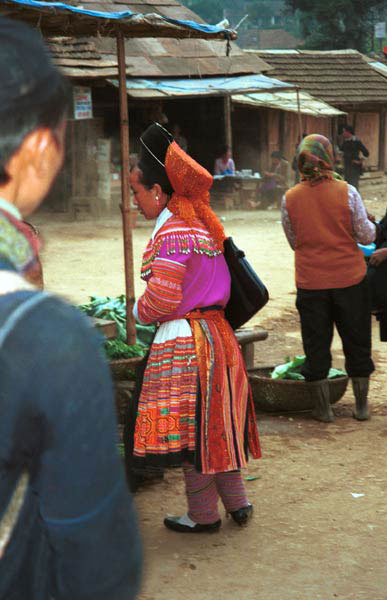 Jpeg 42K 9510K28  Brightly dressed Flowery Hmong woman at a shop stall in Sa Pa, Lao Cai Province.  She is a visitor to Sa Pa, in the town on a training programme, and lives near the Chinese border. The Flowery Hmong near to the border tend to wear more bought trimmings on their clothing which they obtain from China.  Note her embroidered and applique leg wrappings and her long apron.  She only very narrow bands of wax resist on her skirt which features applied strips of fabric and embroidery. 