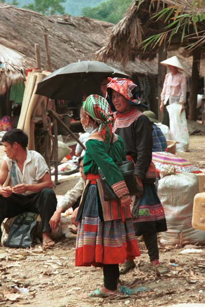 Jpeg 51K  9510K31 Flowery Hmong women at a small local market on the way down from the mountains around Sa Pa towards the town of Lao Cai, Lao Cai Province. Note their plain indigo dyed leg wrappings; mid-length skirt with applique, wax resist insert with some applique; relatively short front and back machine applique aprons; and blouses - one purchased velour and the other indigo dyed woven bast or purchased cotton fabric with applique and possibly some embroidery around the neck, front opening and sleeve bottoms.  On their heads one woman is wearing an imported machine woven woollen scarf from China and the other appears to be wearing an apron to protect her head from the sun.I bought a length of wax resist and a machine applique apron from these women.