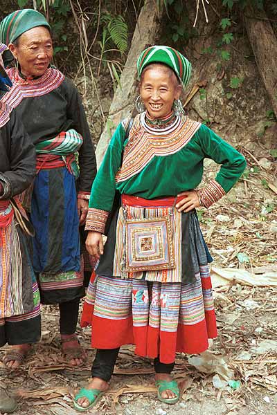 Jpeg 56K  9510K32 Flowery Hmong women at a small local market on the way down from the mountains around Sa Pa towards the town of Lao Cai, Lao Cai Province. Note their plain indigo dyed leg wrappings; mid-length skirts with machine applique, wax resist with some applique; relatively short front and back machine applique aprons; machine applique bag hanging at the waist; and blouses - one purchased velour and the other indigo dyed woven bast or purchased cotton fabric with applique and possibly some embroidery around the neck, front opening and sleeve bottoms.  On their heads both women are wearing imported machine woven woollen scarfs from China. The woman in the centre of the photograph is wearing a white metal necklace and some striking earrings. I bought a length of wax resist and a machine applique apron from these women.