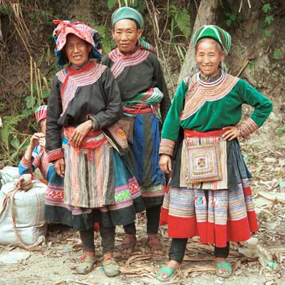 Jpeg 37K 9510K33 Flowery Hmong women at a small local market on the way down from the mountains around Sa Pa towards the town of Lao Cai, Lao Cai Province. Note their plain indigo dyed leg wrappings; mid-length skirts with machine applique, wax resist insert with some applique, embroidered insert; relatively short front and back machine applique aprons; machine applique bags hanging at the waists of two of the women; and blouses - one purchased velour and two indigo dyed woven bast or purchased cotton fabric with applique and possibly some embroidery around the neck, front opening and sleeve bottoms.  On their heads two women are wearing imported machine woven woollen scarfs from China and the other appears to be wearing an apron to protect her head from the sun.I bought a length of wax resist and a machine applique apron from these women.