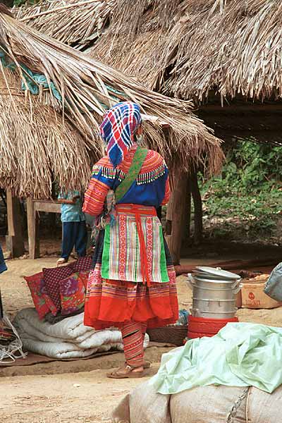 Jpeg 57K  9510K34 Flowery Hmong woman at a small local market on the way down from the mountains around Sa Pa towards the town of Lao Cai, Lao Cai Province.  Her machine applique leg wrappings are similar to the Flowery Hmong woman photographed in Sa Pa who lived near the border with China.  This influence continues in the heavy machine applique and emboridered panels on the sleeves and neck of the blouse as well as the dangling trimmings imported from China.  It is difficult to see whether she has any wax resist inserts on her skirt, although it looks as if possibly the top section is indigo dyed wax resist.  There is a wide red purchased fabric bottom border.  Her apron is relatively short and machine appliqued.