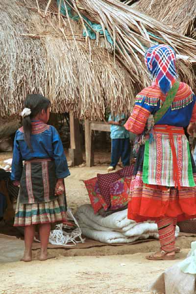 Jpeg 43K  9510K35 Flowery Hmong woman and young girl at a small local market on the way down from the mountains around Sa Pa towards the town of Lao Cai, Lao Cai Province.  the woman's machine applique leg wrappings are similar to the Flowery Hmong woman photographed in Sa Pa who lived near the border with China.  This influence continues in the heavy machine applique and emboridered panels on the sleeves and neck of the blouse as well as the dangling trimmings imported from China.  It is difficult to see whether she has any wax resist inserts on her skirt, although it looks as if possibly the top section is indigo dyed wax resist.  There is a wide red purchased fabric bottom border.  Her apron is relatively short and machine appliqued. The young girl's clothing is a simpler version of the adult clothing.