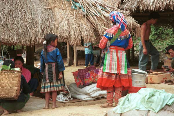 Jpeg 53K  9510K36 Flowery Hmong woman and young girl at a small local market on the way down from the mountains around Sa Pa towards the town of Lao Cai, Lao Cai Province.  the woman's machine applique leg wrappings are similar to the Flowery Hmong woman photographed in Sa Pa who lived near the border with China.  This influence continues in the heavy machine applique and emboridered panels on the sleeves and neck of the blouse as well as the dangling trimmings imported from China.  It is difficult to see whether she has any wax resist inserts on her skirt, although it looks as if possibly the top section is indigo dyed wax resist.  There is a wide red purchased fabric bottom border.  Her apron is relatively short and machine appliqued. The young girl's clothing is a simpler version of the adult clothing.